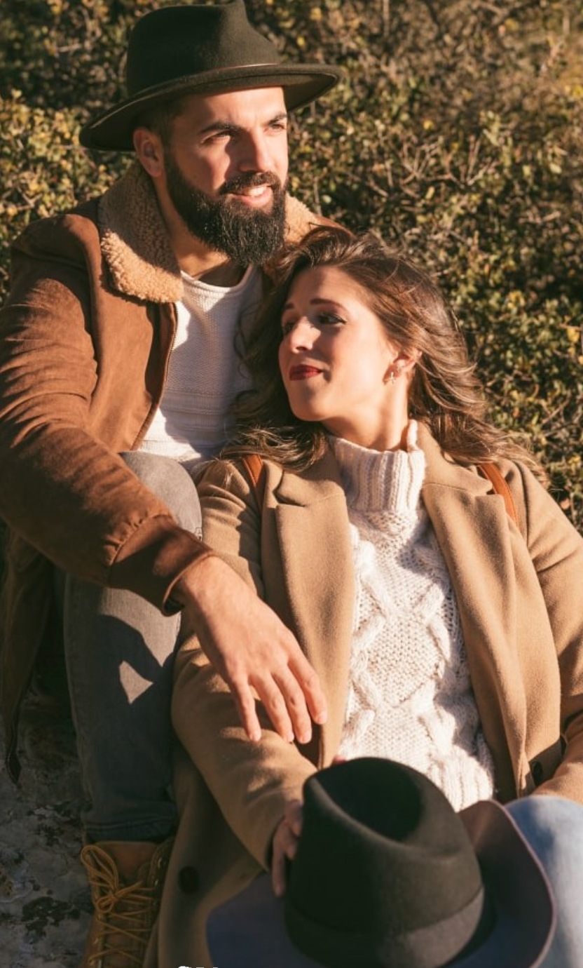 Pareja con abrigos y sombreros sentada al sol junto a unos arbustos